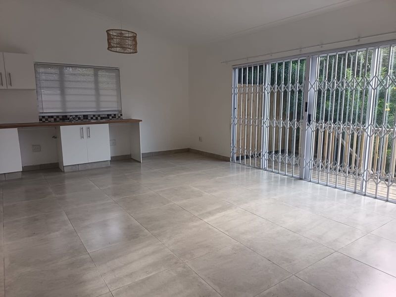 Spacious open-plan living area featuring tiled floors, a compact kitchen kitchenette, and large sliding doors leading to an outdoor space with security gates.