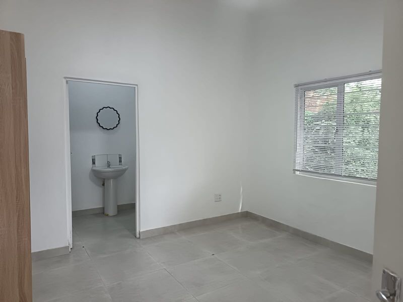 A clean, empty bedroom featuring neutral tiled flooring, large windows with venetian blinds, and an adjoining en-suite area with a pedestal basin.