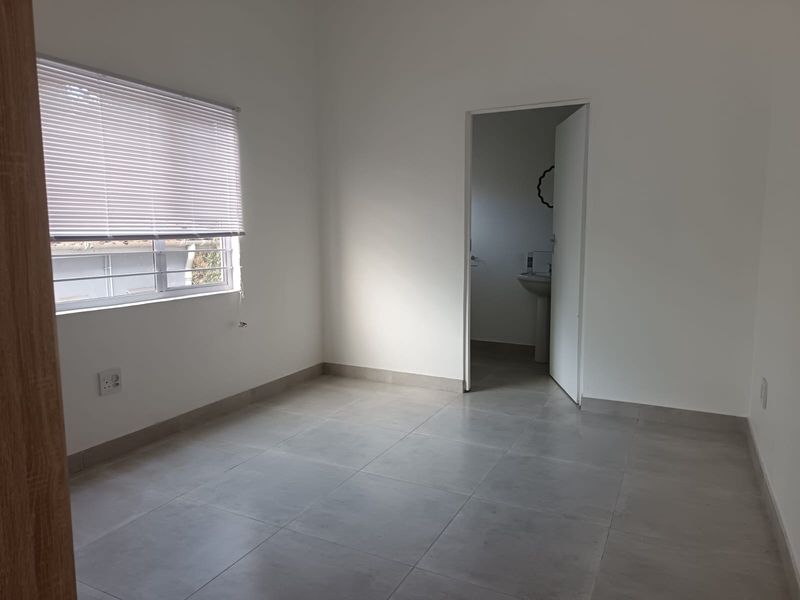 A clean, empty bedroom featuring neutral tiled flooring, white walls, and a window fitted with venetian blinds, with an adjoining bathroom visible.