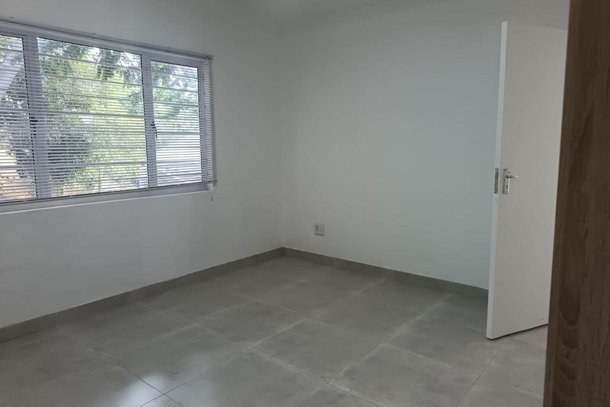 A clean, empty bedroom featuring neutral tiled flooring, white walls, and a window fitted with venetian blinds.