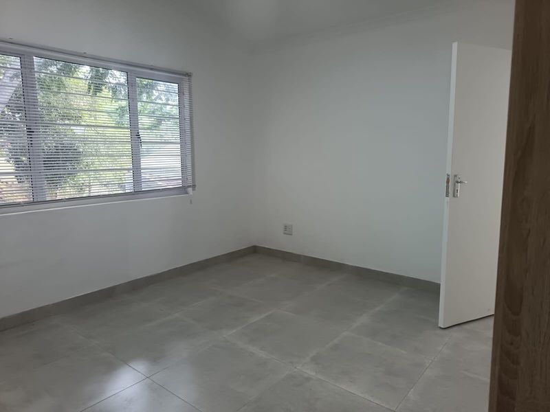 A clean, empty bedroom featuring neutral tiled flooring, white walls, and a window fitted with venetian blinds.