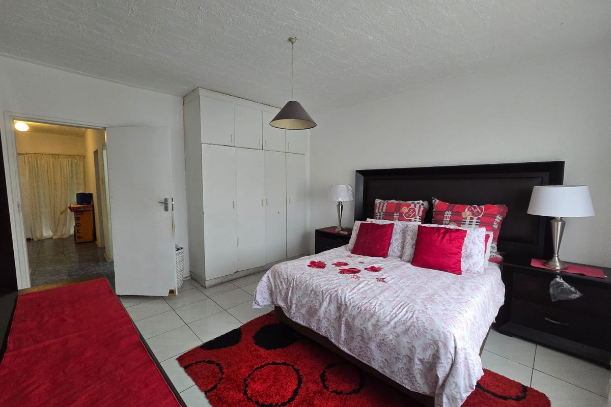 A spacious bedroom featuring built-in cupboards, neutral tiled flooring, and a central pendant light fixture, offering a functional layout with direct access to the hallway.