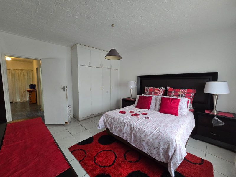 A spacious bedroom featuring built-in cupboards, neutral tiled flooring, and a central pendant light fixture, offering a functional layout with direct access to the hallway.