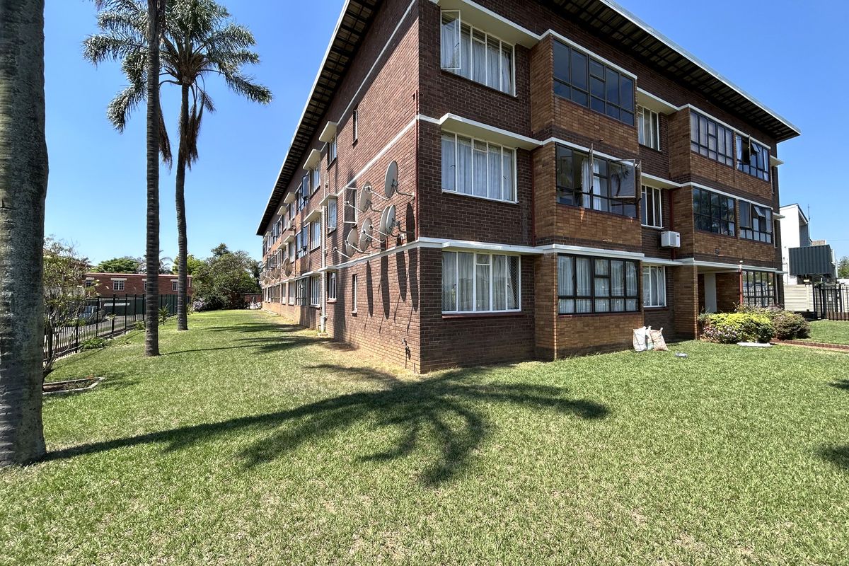 Exterior view of a multi-story brick apartment building featuring metal window frames, multiple satellite dishes, and surrounding communal lawn area.