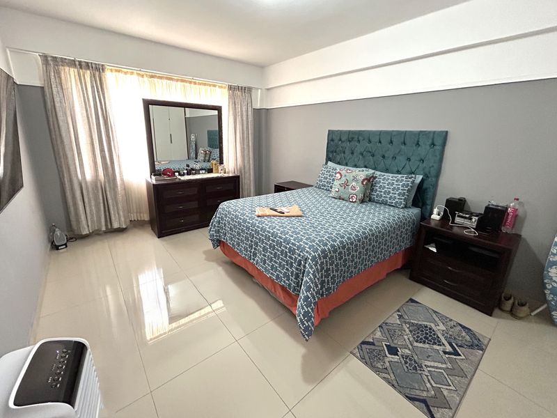 A spacious bedroom featuring neutral grey walls, large windows with curtains, and polished tiled flooring.