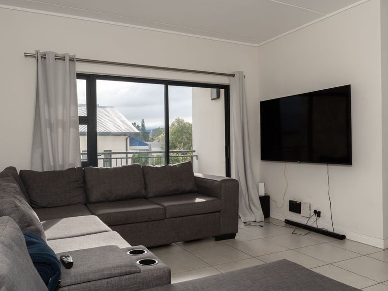 A bright living room featuring neutral tiled flooring, large sliding glass doors providing natural light, and a wall-mounted television setup.