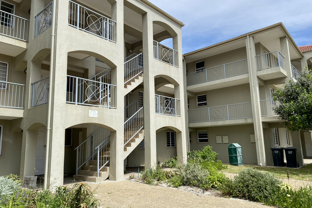 Exterior view of a multi-story apartment complex featuring concrete staircases, open balconies with metal railings, and a paved ground-level walkway.