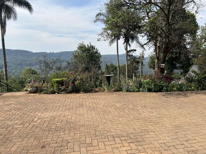 A scenic outdoor view from a brick-paved patio overlooking a lush garden and expansive mountain landscape.