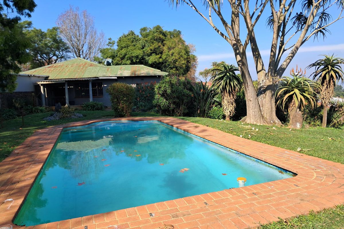 Outdoor pool area featuring a rectangular swimming pool with brick coping, set within a garden with mature trees and a house with a metal roof.