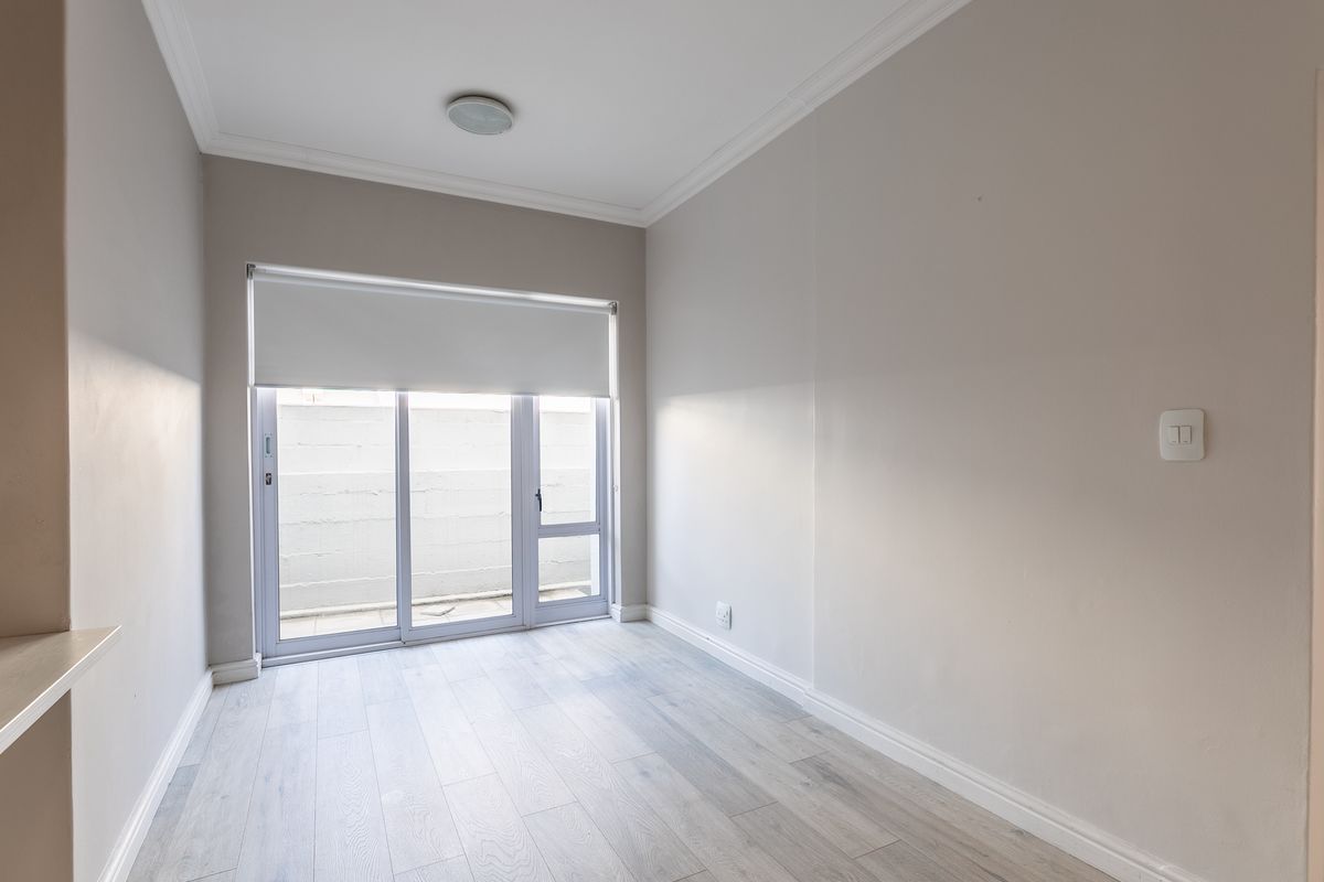 A bright, empty bedroom featuring light-toned laminate flooring, neutral walls, and a sliding glass door with a roller blind leading to an outdoor area.