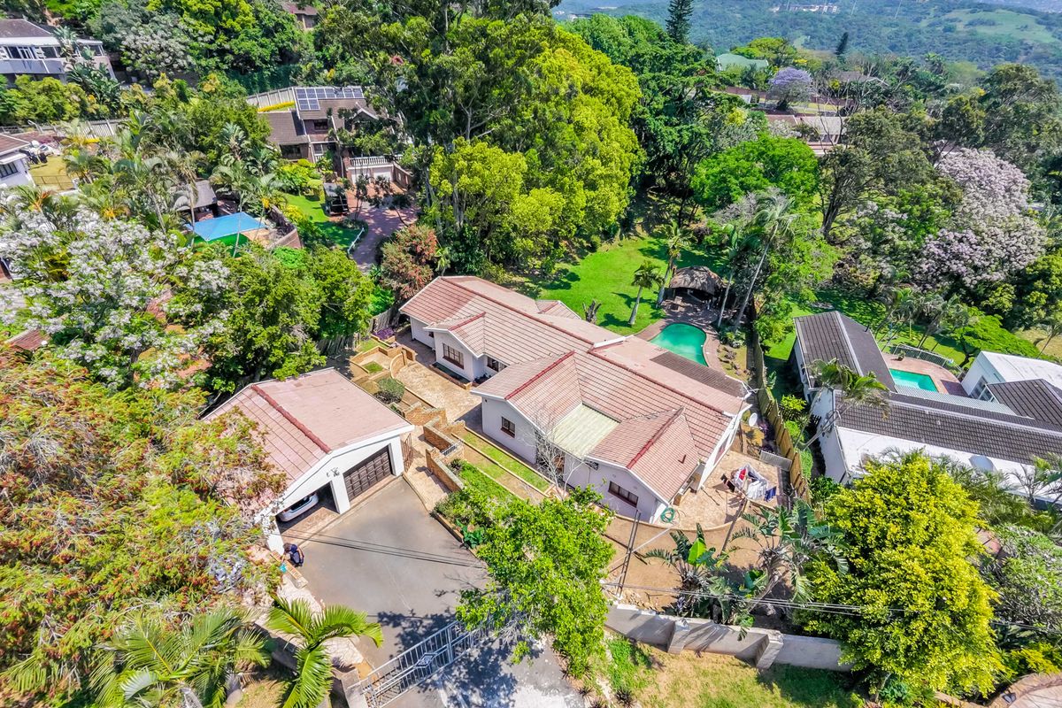 Aerial view of a residential property featuring a main house, detached garage, swimming pool, and established garden within a lush, green suburban setting.
