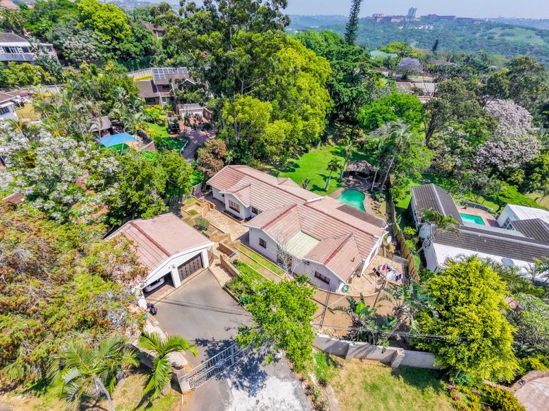 Aerial view of a residential property featuring a main house, detached garage, swimming pool, and established garden within a lush, green suburban setting.