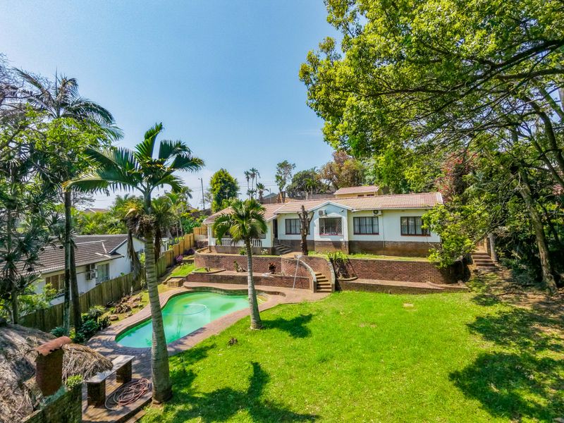 Rear view of the property featuring a swimming pool, tiered brick retaining walls, and a spacious garden area with mature trees.