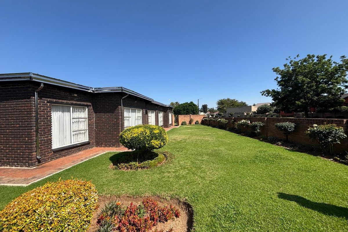 A well-maintained rear garden featuring a spacious lawn, brick boundary wall, and a paved walkway along the dark brick house exterior.