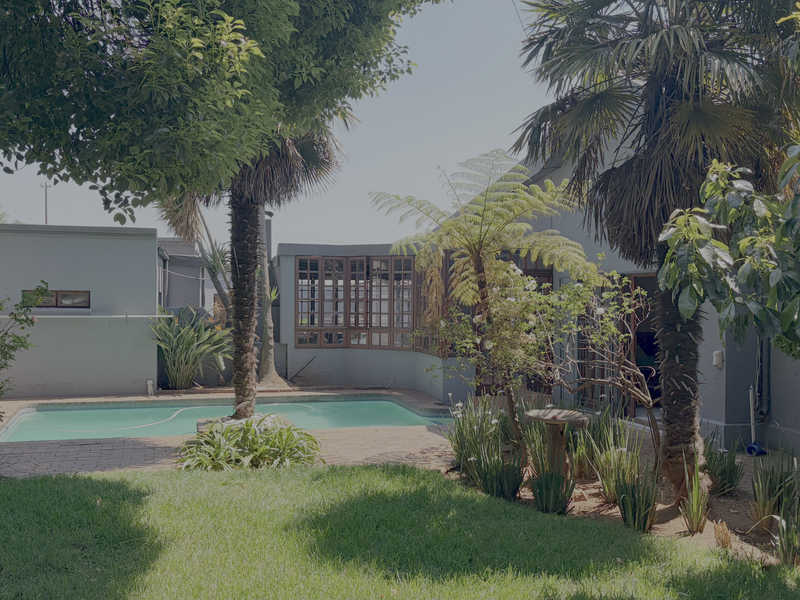 A private backyard featuring a swimming pool, brick-paved patio area, and established garden landscaping with mature trees and shrubs.