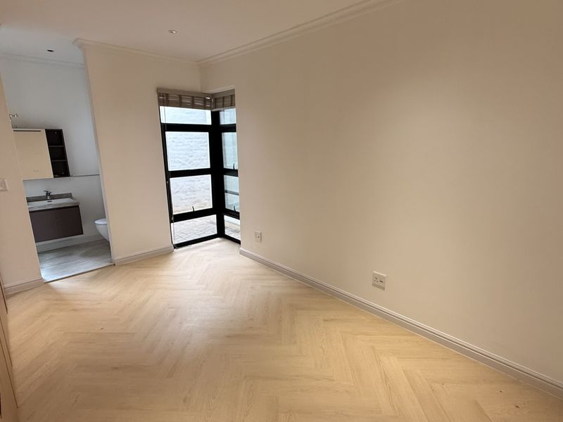 A bright, empty bedroom featuring modern herringbone timber flooring, built-in cabinetry, and an adjoining en-suite bathroom with a corner window.