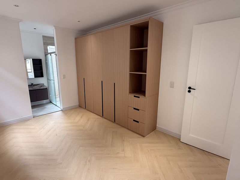 A bright, modern bedroom featuring light wood herringbone flooring, a large built-in wardrobe with integrated shelving, and direct access to an en-suite bathroom.