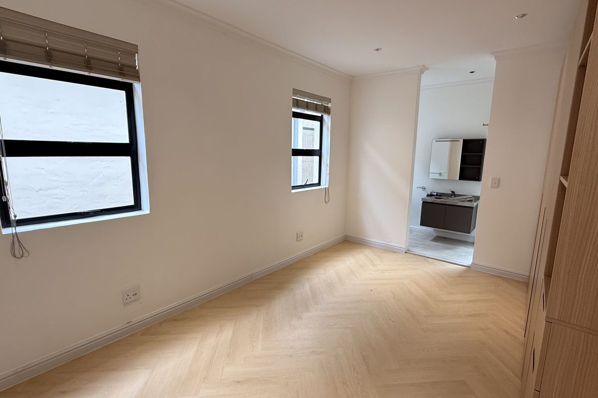 A bright, empty bedroom featuring modern herringbone flooring, built-in cupboards, and an adjoining en-suite bathroom with neutral finishes.