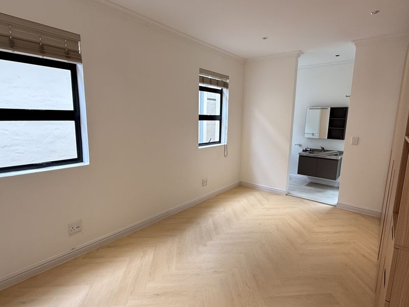 A bright, empty bedroom featuring modern herringbone flooring, built-in cupboards, and an adjoining en-suite bathroom with neutral finishes.