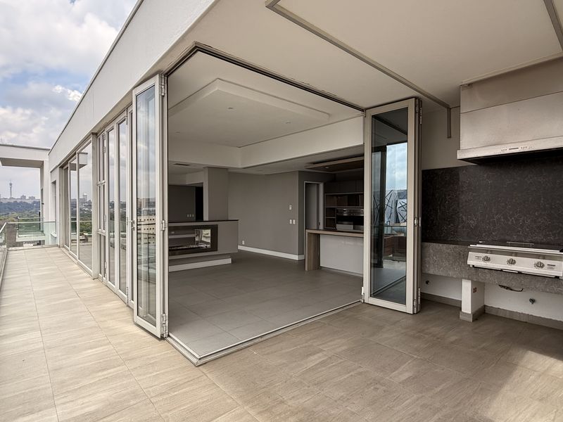 Spacious penthouse balcony featuring a built-in gas braai, stone splashback, glass balustrades, and seamless indoor-outdoor flow through large bi-fold doors.