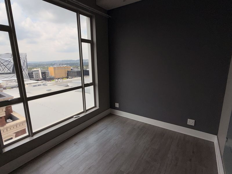 A bright, empty bedroom featuring modern laminate flooring, dark accent walls, and expansive floor-to-ceiling windows offering an urban city view.