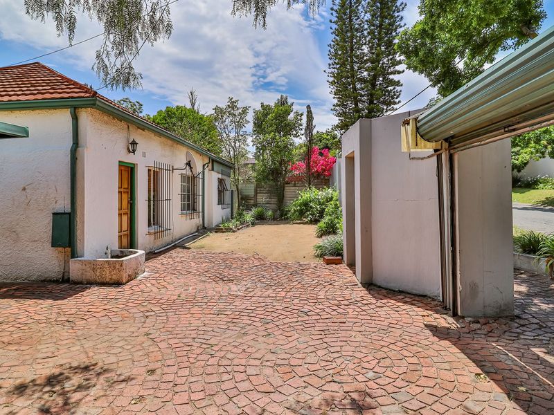 Rear exterior view of the property featuring a brick-paved courtyard, carport structure, and established garden area with security features.