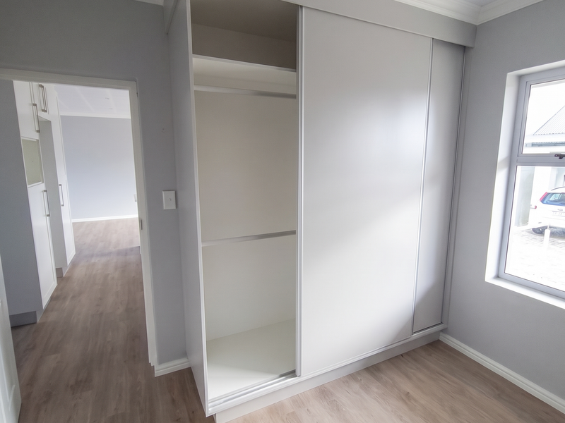 A bright, modern bedroom featuring light laminate flooring, a built-in sliding wardrobe, and a clean, neutral aesthetic with crown molding details.