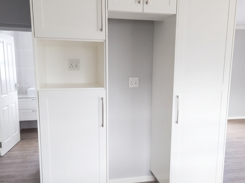 A modern, clean kitchen area featuring white built-in cabinetry with dedicated appliance housing and integrated electrical points, set against light-toned laminate flooring.
