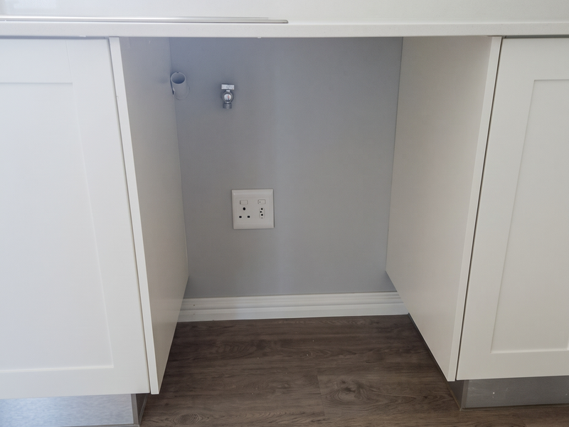 A clean, modern laundry appliance recess featuring white cabinetry, stone countertop, and dedicated plumbing and electrical connections for a washing machine.