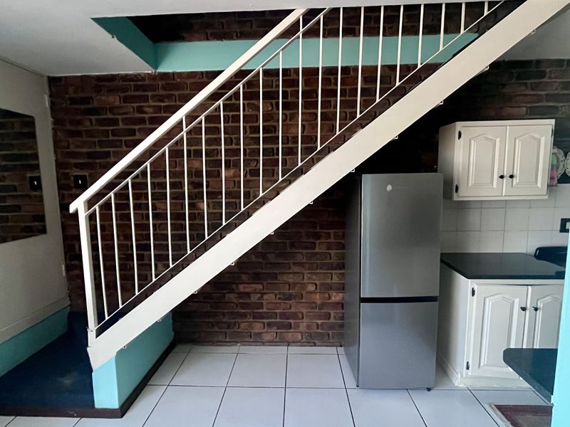 Open plan area featuring a staircase with metal balustrade, exposed brick wall, tiled flooring, and built-in kitchen cabinetry with dark countertops.