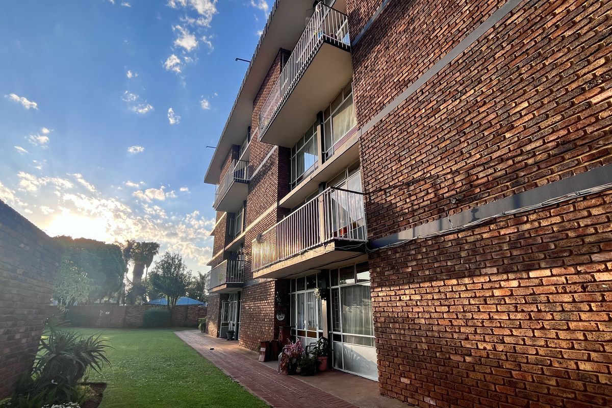A view of the rear exterior of a multi-story apartment building featuring exposed brickwork, private balconies with metal railings, and a shared garden area.