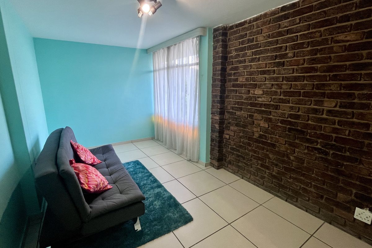 A living room featuring a prominent exposed brick feature wall, light blue painted walls, and neutral tiled flooring with natural light from the window.