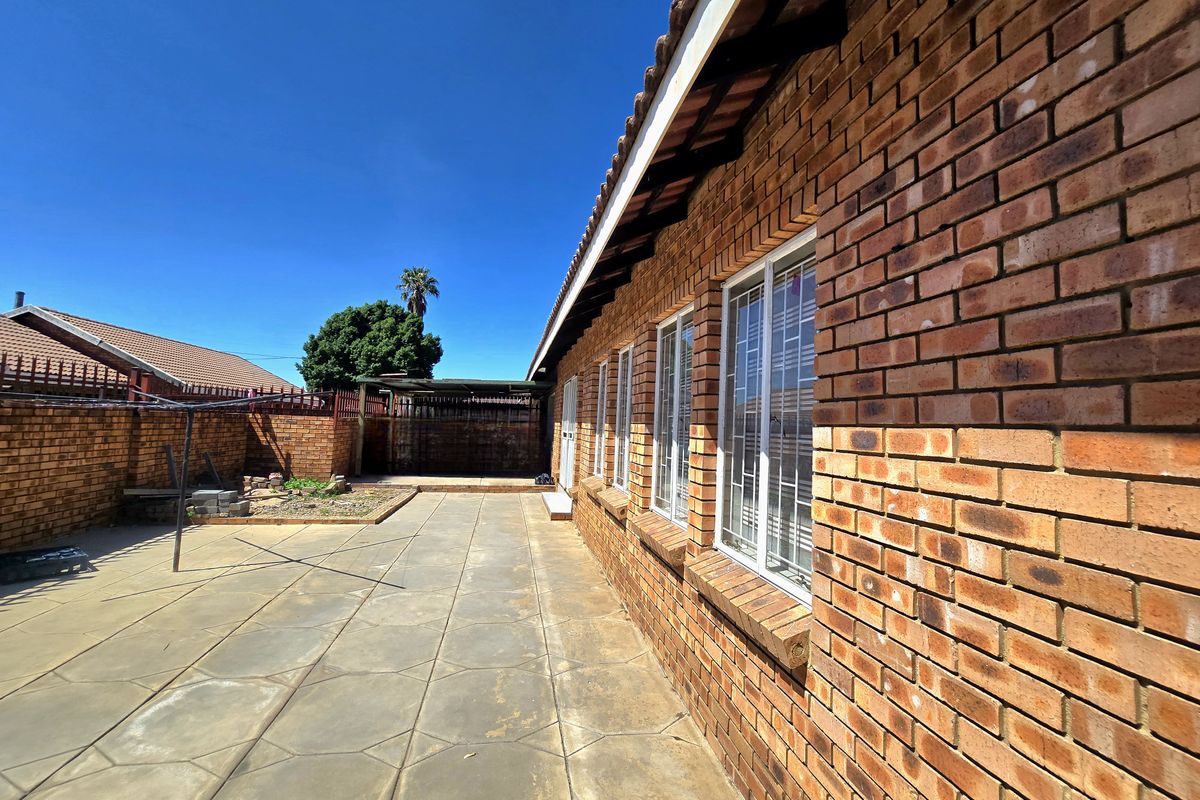 A side view of the townhouse featuring face brick exterior walls, a paved patio area, and secure windows with fitted security bars.
