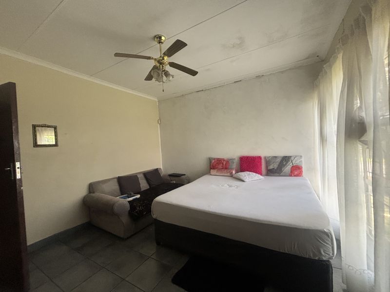 A bedroom featuring tiled flooring, a ceiling fan, and large windows. The room requires maintenance due to visible ceiling and wall finish deterioration.