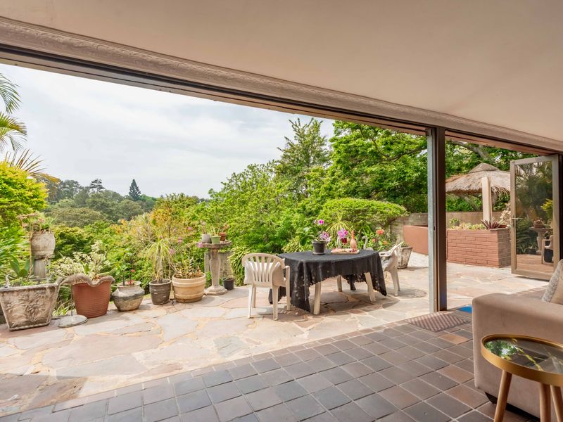 A spacious covered patio featuring stone and tile flooring, seamlessly connecting to a lush garden area through large sliding glass doors.