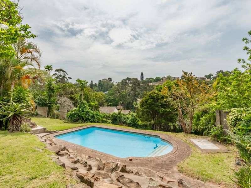 A private swimming pool set within a lush, established garden, featuring brick coping and a scenic outlook over the surrounding neighborhood.