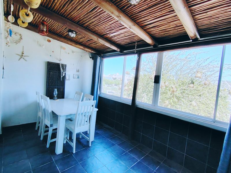 A covered patio area featuring a rustic thatched roof with exposed timber beams, tiled flooring, and large sliding windows overlooking the garden.