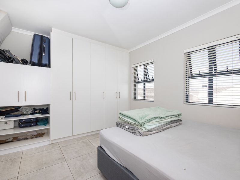 A bright bedroom featuring neutral tiled flooring, ample built-in cupboard storage, and large windows with fitted venetian blinds.