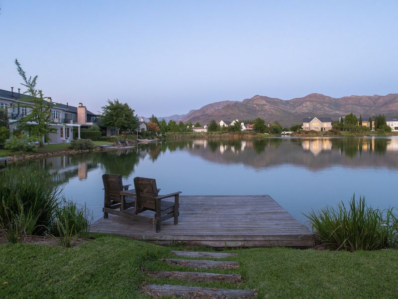 A scenic view of a tranquil lake with a wooden jetty, surrounded by landscaped gardens and a mountain backdrop.