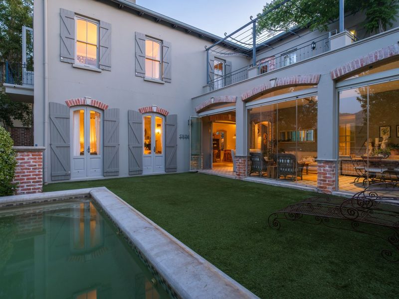 A beautifully maintained rear exterior featuring a swimming pool, manicured lawn, and elegant architectural details including brick accents and glass folding doors.