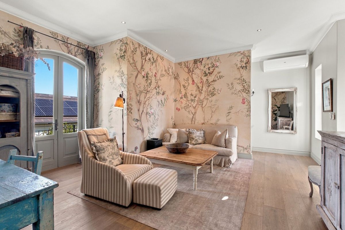 A bright and elegant living room featuring timber flooring, decorative floral wallpaper, recessed lighting, and arched french doors leading to an outdoor area.