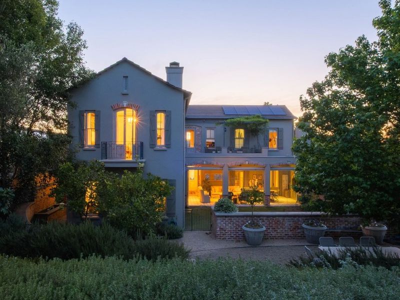 A beautiful rear elevation of a two-story home featuring a covered patio, balcony, solar panels, and a landscaped garden at dusk.