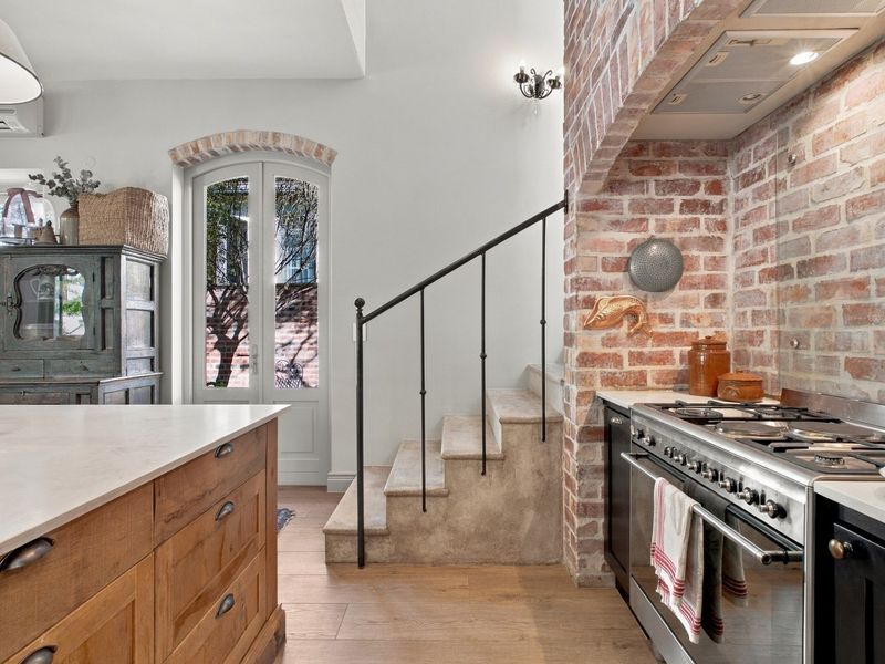 A characterful kitchen featuring an exposed brick wall, professional-style gas range, stone staircase with iron railing, and direct access to the garden.