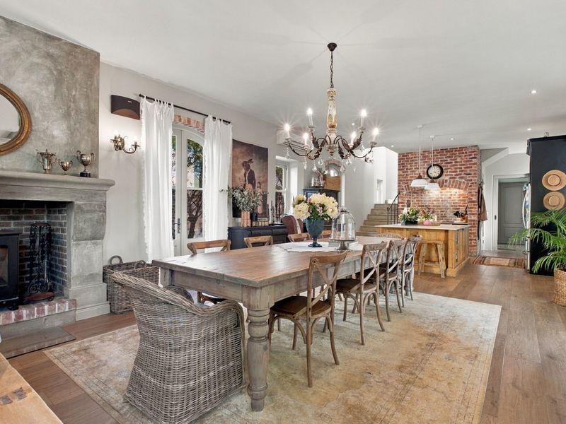 Spacious open-plan living and dining area featuring a stone fireplace, wood-burning stove, exposed brick accents, and a central kitchen island with hardwood flooring.