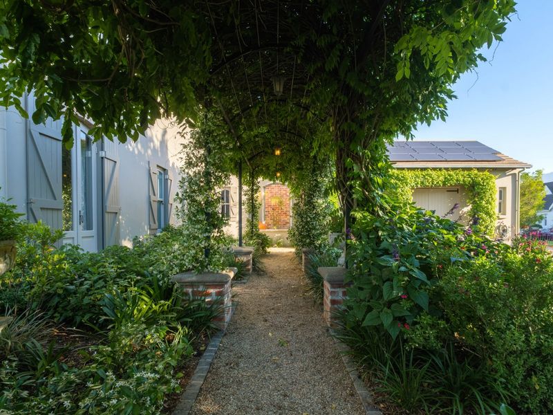 A beautifully landscaped garden path featuring a vine-covered metal pergola, brick pillars, and established greenery leading alongside the property.
