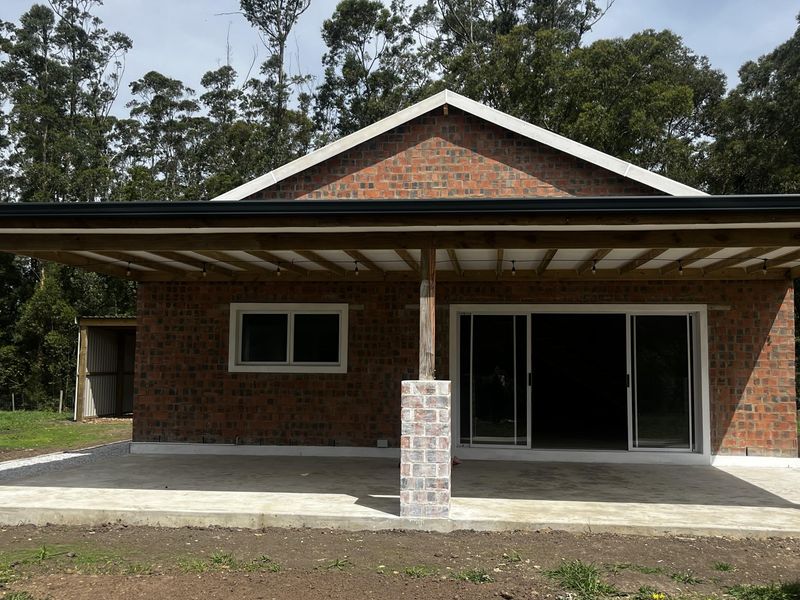 A covered outdoor patio area featuring exposed brickwork, a concrete slab, and sliding glass doors, set against a natural rural backdrop.