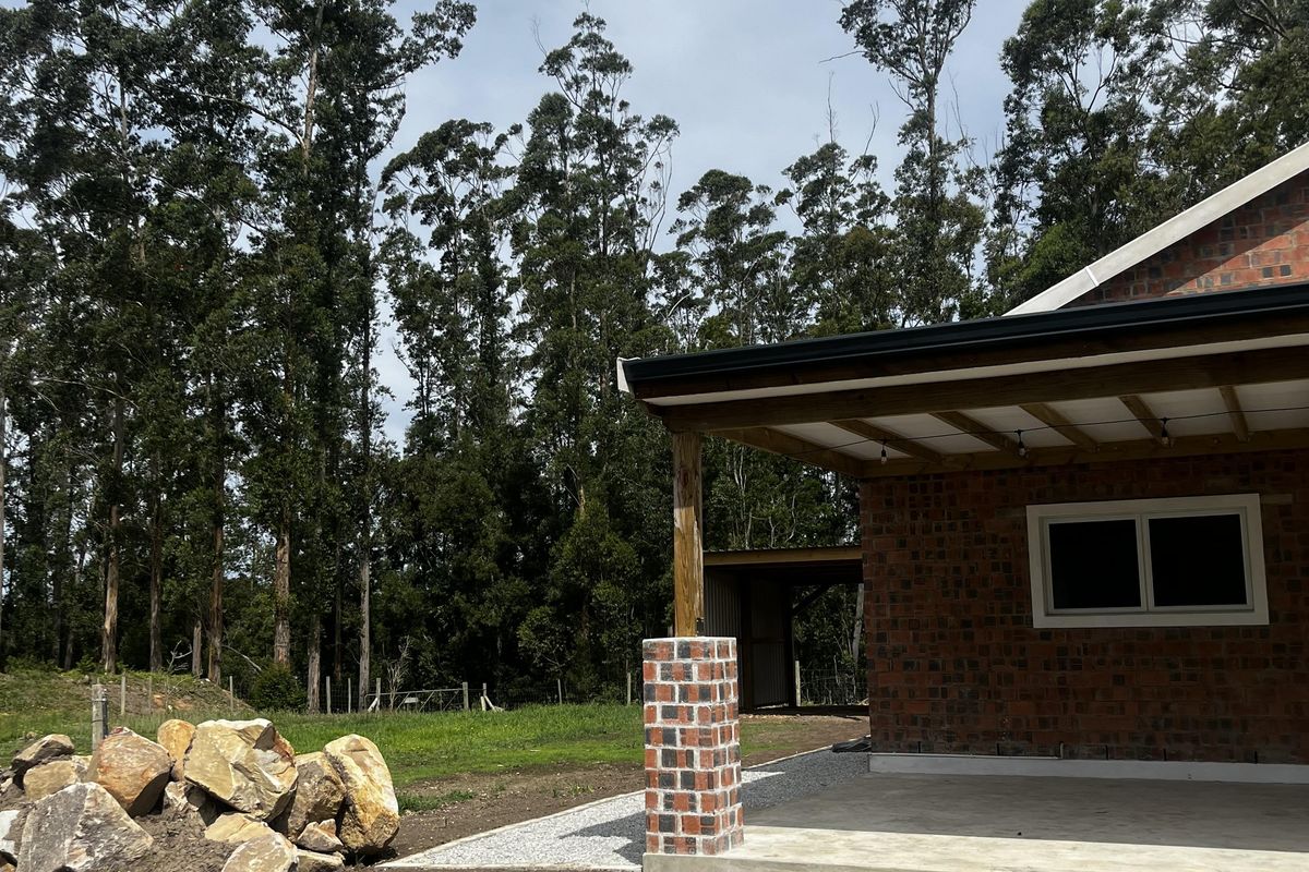 A rear view of a farm property featuring an exposed brick exterior, a concrete patio area, and a gravel pathway surrounded by mature trees.