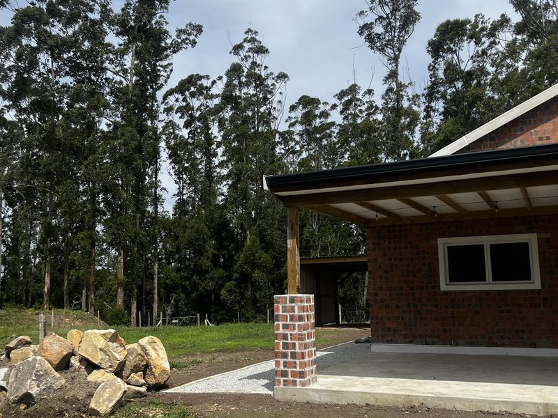A rear view of a farm property featuring an exposed brick exterior, a concrete patio area, and a gravel pathway surrounded by mature trees.