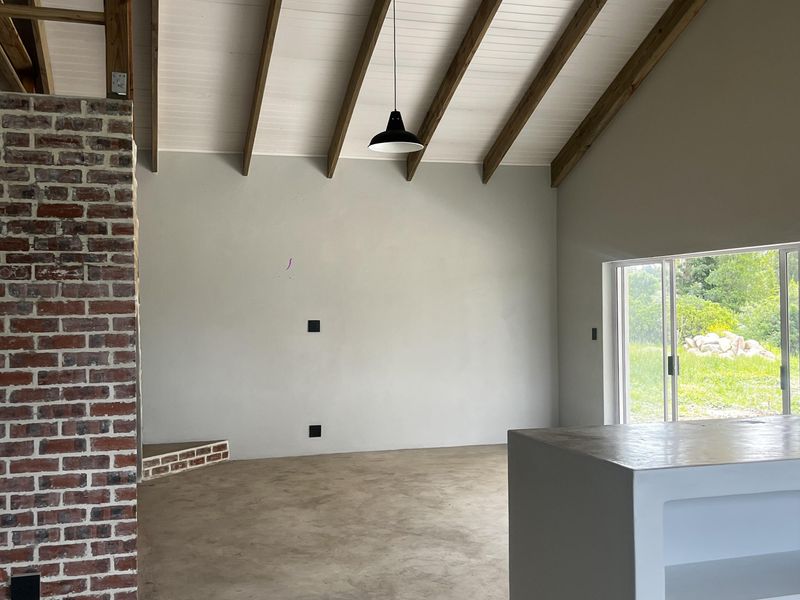 Spacious open-plan living area featuring exposed timber beams, a striking brick feature wall, polished concrete floors, and direct access to the garden.