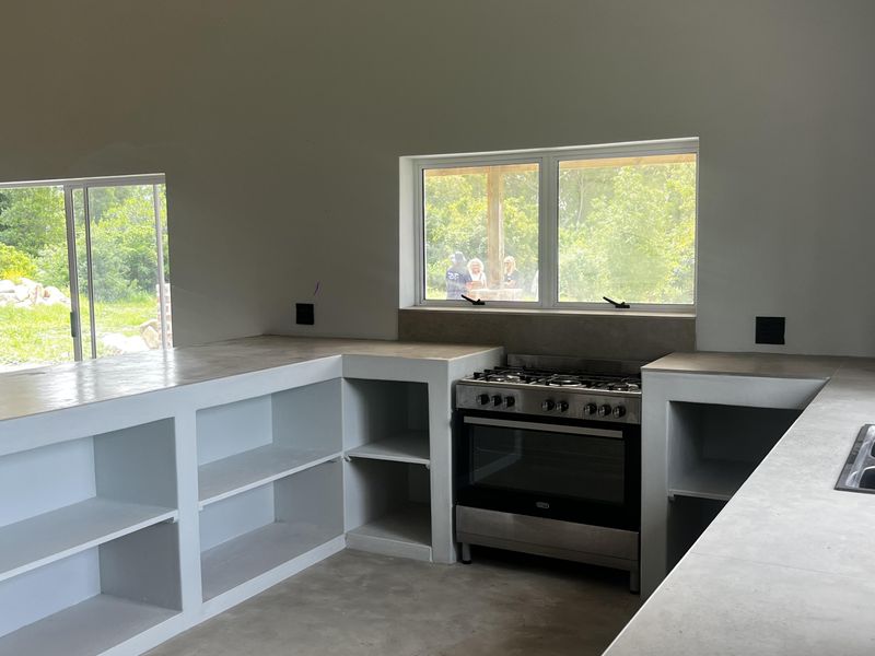 A modern, minimalist kitchen featuring custom built-in concrete shelving, concrete countertops, and a stainless steel gas stove, set within a bright, open-plan space.